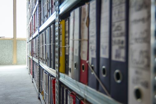 Binders and folders with archive documents are lined up along shelves in a storage room.